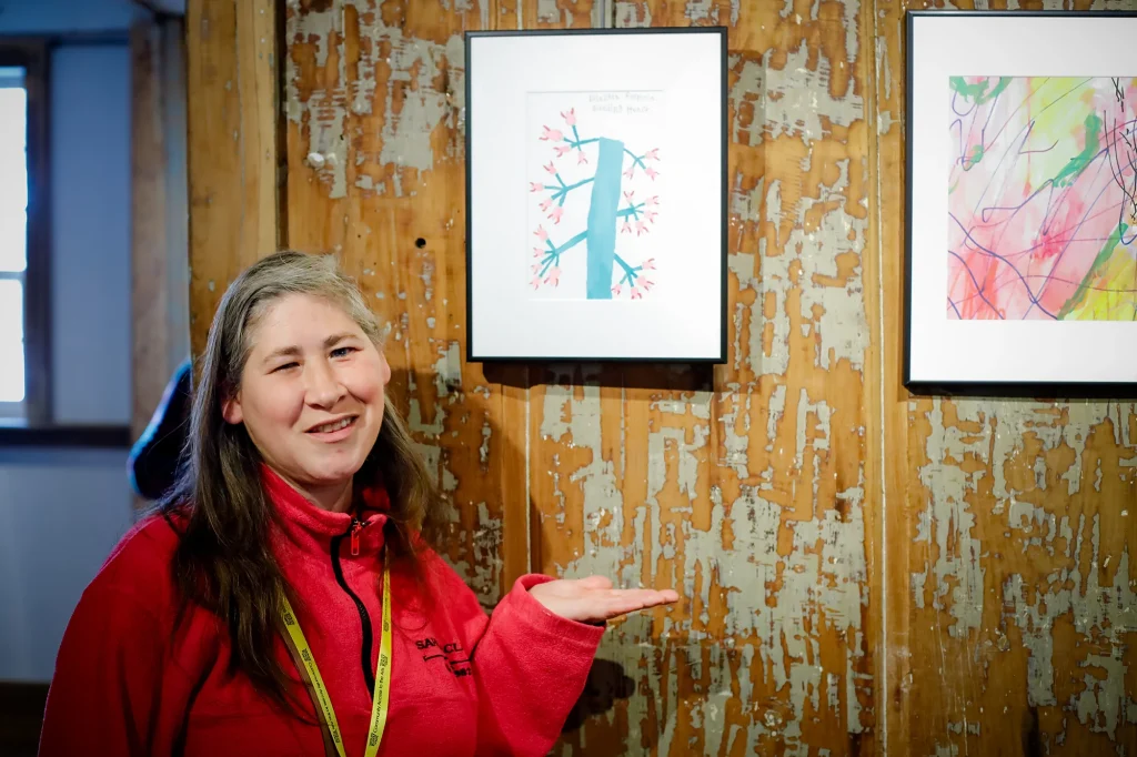 A woman stands gesturing to artwork in a gallery.