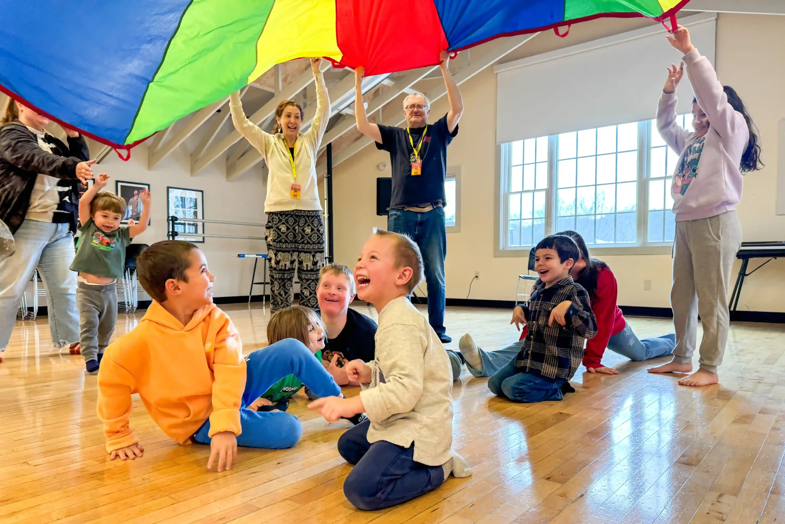 A group of children laugh while crouching beneath a colorful parachute.