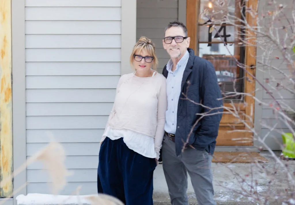 Two people wearing glasses and warm clothes smile in front of a house in the winter.